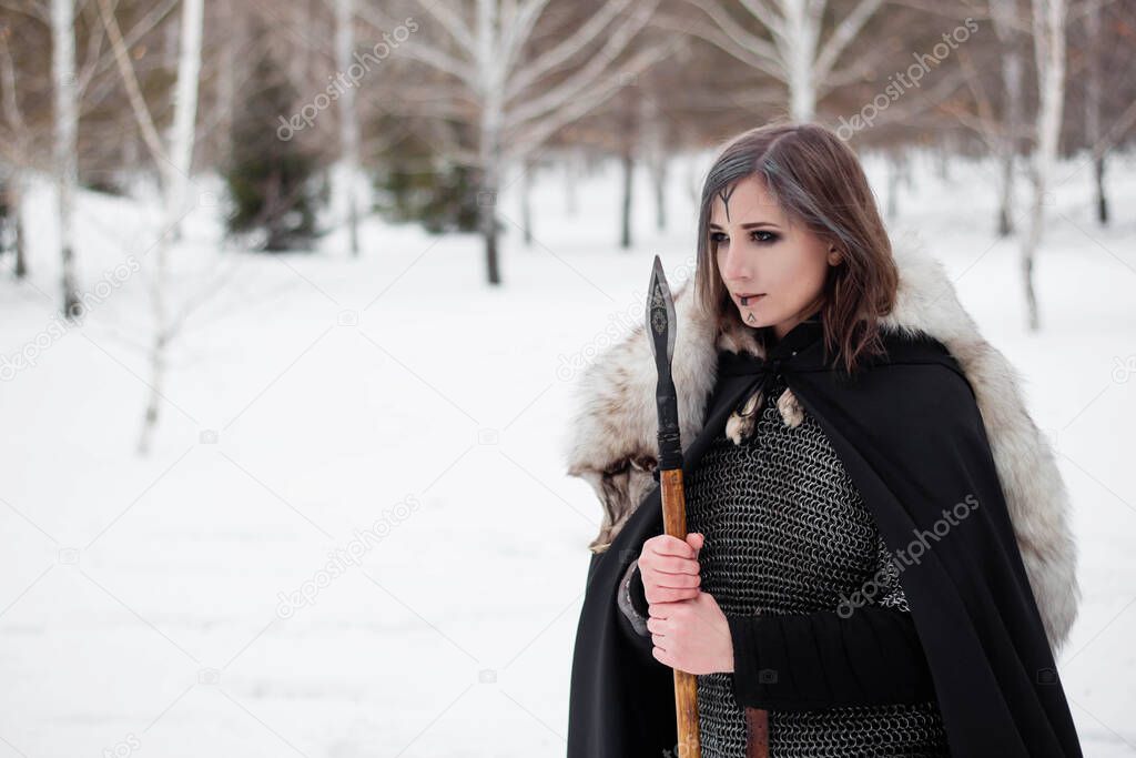 The woman is a medieval warrior of the Viking Age in the winter in the forest. Dressed in chain mail, cloak, fur,  leather pants and boots with a spear in their hands. Battle coloring on the face.