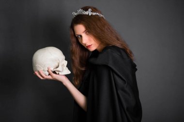 A beautiful, young woman with long hair in a black cloak with a hood and diadem on her head in her hands holds a skull. Studio photo on a gray background. Witchcraft, necromancy, divination, Halloween. clipart