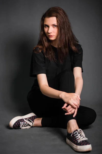 美しい 長い髪を持つ若い女性は 黒い服の床に座っている グレーの背景にスタジオ写真 きれいな肌をしたモデル — ストック写真