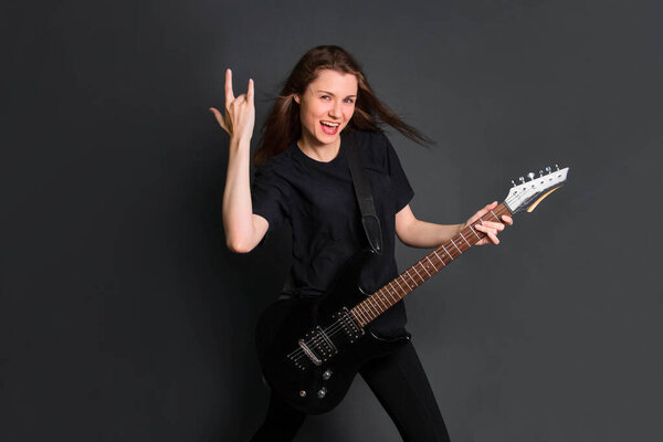 Красивая молодая рок-женщина в черной одежде с электрогитарой в руках показывает козлиный жест. Студийное фото на сером фоне. Модель с чистой кожей
.
