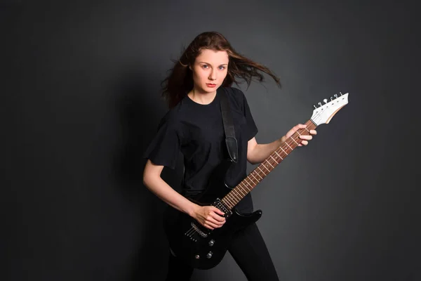 Portrait of a beautiful, young rock woman in black clothes with an electric guitar in her hands. Studio photo on a gray background. Model with clean skin.