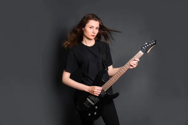 Portrait of a beautiful, young rock woman in black clothes with an electric guitar in her hands. Studio photo on a gray background. Model with clean skin.