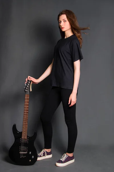 Attractive, young rock woman in black clothes with an electric guitar in her hands stands on a gray background. Model with clean skin.