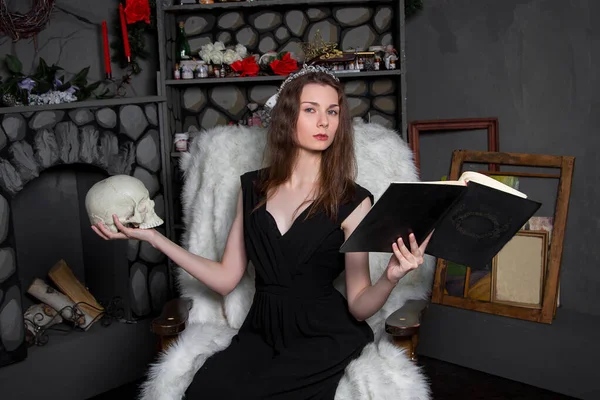 頭にシルバーのダイヤモンドが施された黒いドレスを着た若い美しい女性が 頭蓋骨と手に本を持って暖炉の近くのロッキングチェアに座っています 彼は呪文を読み 思いを馳せる ハロウィン — ストック写真