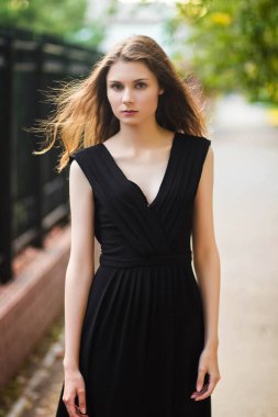 Siyah elbiseli genç, güzel bir kadın. Çelik çitlerin arkasında, açık hava modasına uygun bir fotoğraf. Yaz akşamı. Temiz ciltli manken.