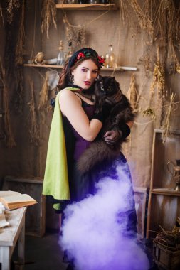 Elinde siyah bir kedi olan genç ve çekici bir cadı sihirli mor duman yayan büyük siyah bir kazanda iksir pişiriyor..
