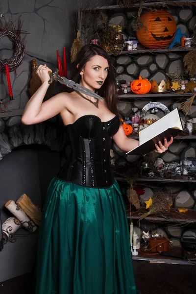 一个穿着绿色裙子和黑色紧身衣的年轻漂亮姑娘手里拿着一本书和一把刀 万圣节的女巫服装 皮肤洁白的模特 — 图库照片