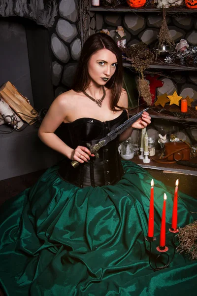一个穿着绿色裙子和黑色紧身衣的年轻漂亮的女人坐在地板上 手里拿着一把点燃的蜡烛 手里拿着一把刀 女巫会变魔术的万圣节 — 图库照片