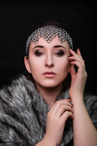 Beauty portrait of a woman with a chain mail bezel on her head and fur on her shoulders. Studio photo on a black background. Model with clear skin, retouch.