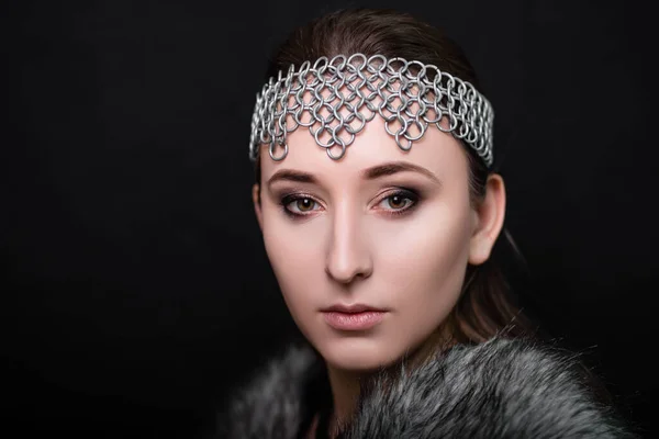 Beauty portrait of a woman with a chain mail bezel on her head and fur on her shoulders. Studio photo on a black background. Model with clear skin, retouch.