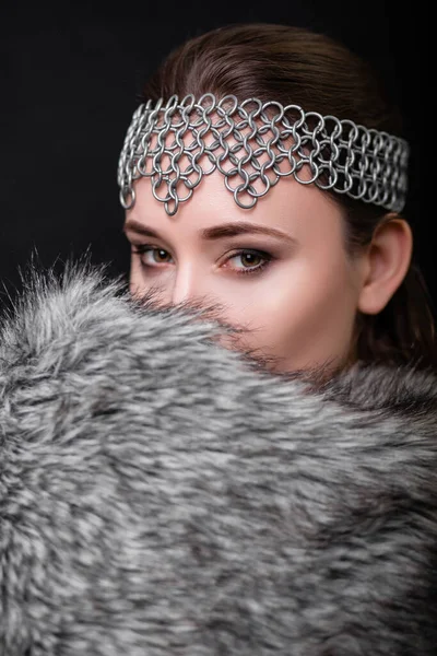 Beauty portrait of a woman with a chain mail bezel on her head and fur on her shoulders. Studio photo on a black background. Model with clear skin, retouch.