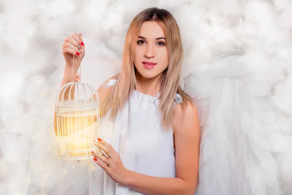 A young girl in a white tunic with large white wings behind her back stands in white clouds and holds a lamp cage with magic light in her hands.