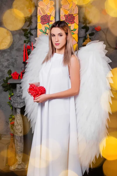 天使は赤いハートを手に持っている 中世の窓を背に白い翼を持つ白いドレスの少女の肖像画と前景に黄色のボケ — ストック写真