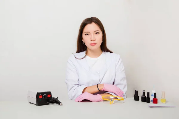 The woman is a master of nails, in a white coat and gloves, sitting at a table with spread manicure tools. Photo on a white background.