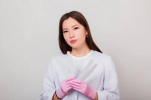 Portrait of a woman, master of nails in a white coat and gloves, standing on a white background with nail files in her hands.