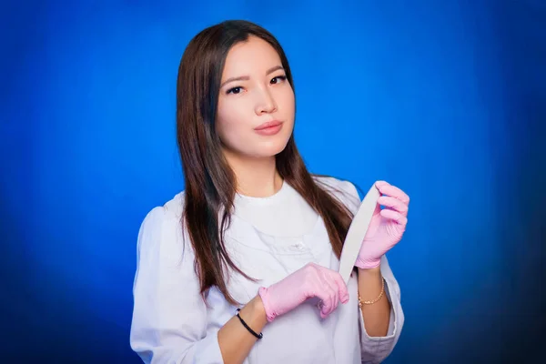 Portrait of a woman master of nails, in a white coat and pink mask on her face with nail files in her hands. Photo on a blue background.