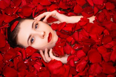 Üst manzara, kırmızı gül yapraklarıyla dolu bir küvette genç bir kadının yakın çekim yüzü. Çekici bir kızın güzel portresi. Cilt yenilenmesi için spa tedavisi.