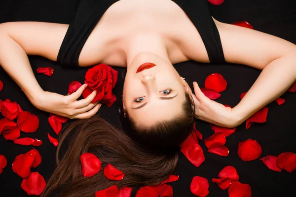 Portrait of a young, attractive woman with a rose in her hands lying on rose petals on a black background. Valentine\'s day concept. Model with retouched skin.