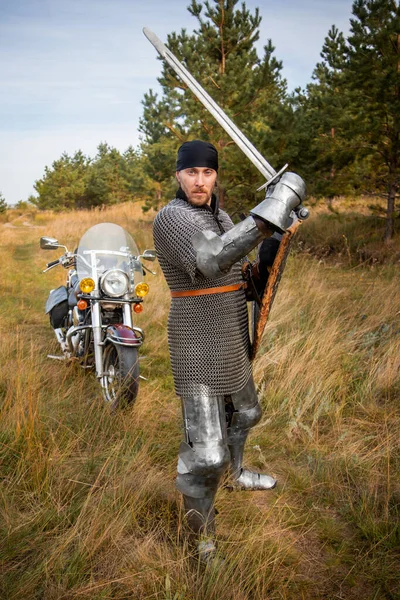 A medieval knight in chainmail with a shield and a sword in his hands stands against the backdrop of a motorcycle and a forest.