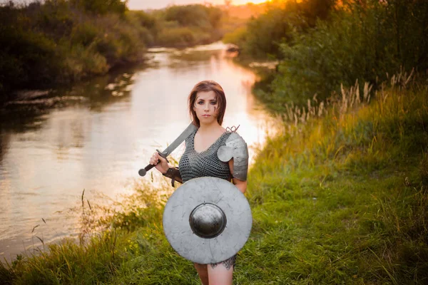 Medieval fantasy female warrior with war paint on her face, in chain mail armor, with a sword and shield in her hands, poses standing against the backdrop of a sunset, river and forest.