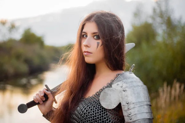 Portrait of a medieval woman warrior in chain mail with a sword and shield in her hands against the backdrop of the river and sunset. Combat makeup, clear skin, retouching.