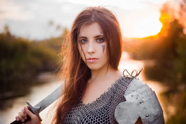 Portrait of a medieval woman warrior in chain mail with a sword and shield in her hands against the backdrop of the river and sunset. Combat makeup, clear skin, retouching.