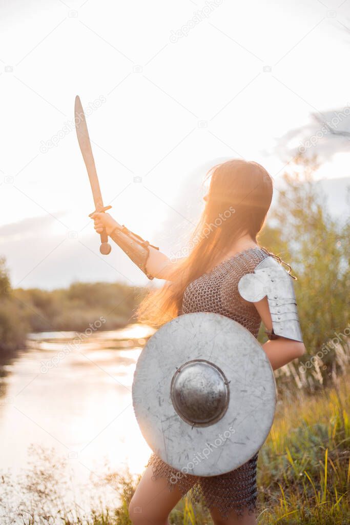 Medieval fantasy female warrior with war paint on her face, in chain mail armor, with a sword and shield in her hands, poses standing against the backdrop of a sunset, river and forest.