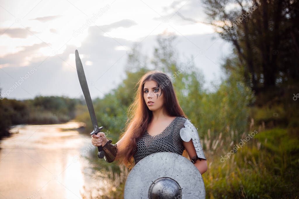 Portrait of a medieval woman warrior in chain mail with a sword and shield in her hands against the backdrop of the river and sunset. Combat makeup, clear skin, retouching.