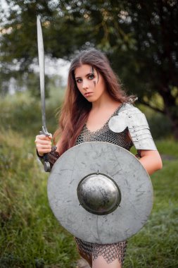 Çatışma makyajlı, zırhlı, plaka vatkalı ve askılı bir kadın savaşçı ormanın arka planına karşı savaş, koruma ve eskrim pozisyonunda poz verir..