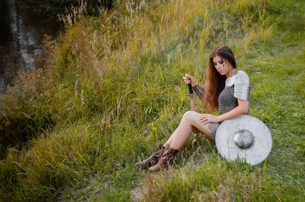 A woman warrior in medieval fantasy armor sits on the grass with a sword in her hands.