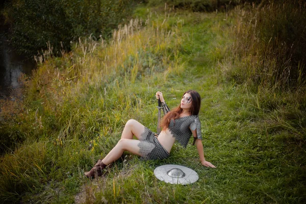 A woman warrior in medieval fantasy armor sits on the grass with a sword in her hands.