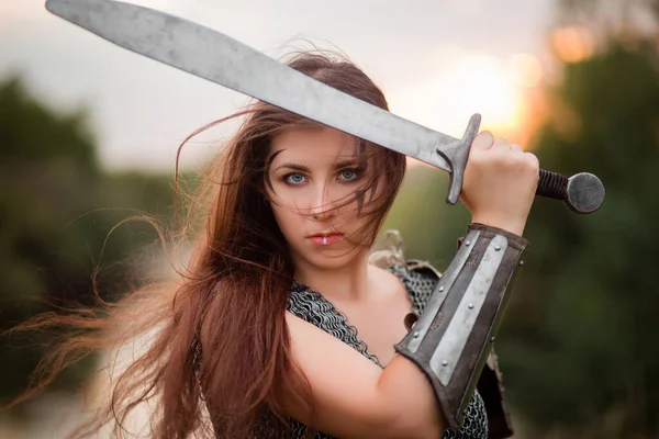 A woman warrior with combat makeup in a chainmail top with plate shoulder pads and bracers poses in a combat, protective, fencing position against the background of the forest.