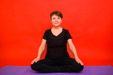 Siyah elbiseli yaşlı bir kadın mor halıda lotus pozisyonunda yoga yapıyor. Kırmızı arkaplanda stüdyo fotoğrafı. 