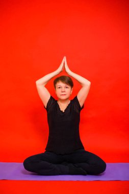 Siyah elbiseli yaşlı bir kadın mor halıda lotus pozisyonunda yoga yapıyor. Kırmızı arkaplanda stüdyo fotoğrafı. 