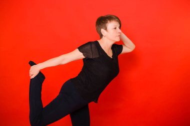 Siyah elbiseli yaşlı bir kadın tek ayak üstünde yoga yapıyor. Kırmızı arkaplanda stüdyo fotoğrafı. 