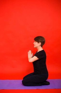 Siyah elbiseli yaşlı bir kadın mor bir halıya oturmuş yoga yapıyor. Kırmızı arkaplanda stüdyo fotoğrafı. 