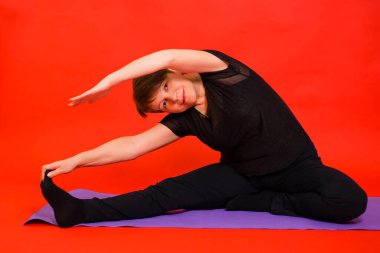 Siyah elbiseli yaşlı bir kadın yoga yapıyor ve mor bir halıda otururken bacaklarını esnetiyor. Kırmızı arkaplanda stüdyo fotoğrafı. 