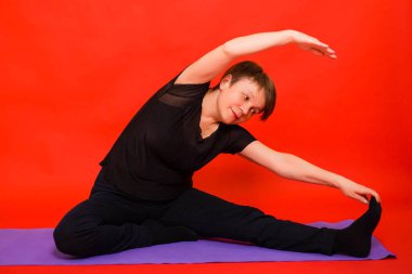 Siyah elbiseli yaşlı bir kadın yoga yapıyor ve mor bir halıda otururken bacaklarını esnetiyor. Kırmızı arkaplanda stüdyo fotoğrafı. 