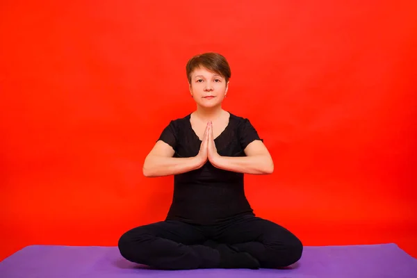 Aged woman in black clothes doing yoga sitting in lotus position on a purple rug. Studio photo on a red background.