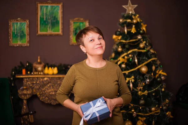 クリスマスのインテリアやクリスマスツリーの背景に立っている間 彼女の手の中に贈り物を保持笑顔高齢女性 — ストック写真