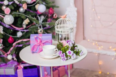 Kutusunda bir hediye olan beyaz masa, çiçekler, bir kafes ve bir fincan tabağın üzerinde bir Noel ağacı ve şöminenin arka planında.
