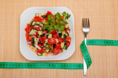 Bir çatal ve bir santimetre, salatalık, domates ve soğan salatasının yanında, beyaz bir tabakta ayçiçeği yağı serpiştirilmiş. Düzgün beslenme konsepti, kilo vermek için diyet..