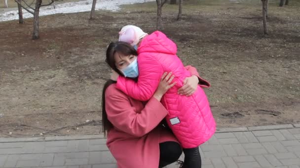 小さな女の子は母親を抱きかかえ春の医療マスクの家族はコロナウイルスのパンデミックの間公園を歩く — ストック動画