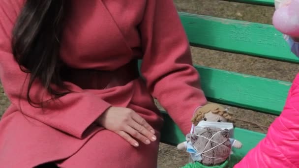 早春的时候 身穿保暖衣服和防护面具的年轻妈妈和女儿坐在公园的长椅上 玩着带医疗面具的软软玩具 — 图库视频影像