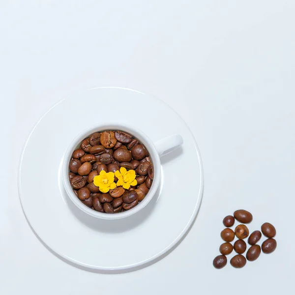 white cup and saucer on a white background top view. in a cup are poured coffee beans and yellow flowers. on a white background heart of coffee beans