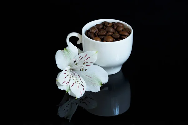 White cup with grains of coffee on a black background. there is a white flower near the cup