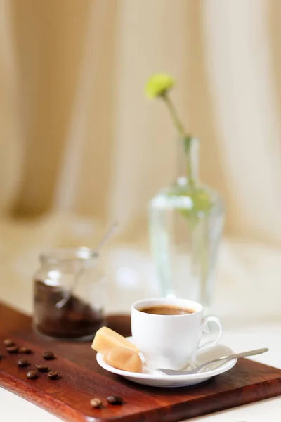 White cup of coffee. Cup and saucer stand on the board. On a saucer are sweets and a coffee spoon. Coffee grains are scattered on the board. In the background is a jar of coffee and a beautiful flower in a vase