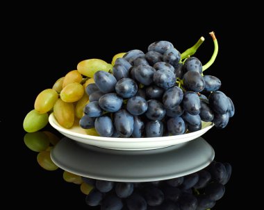 Bir demet yeşil ve mavi üzüm beyaz bir tabakta yatıyor. Plaka siyah parlak arka plan üzerinde duruyor