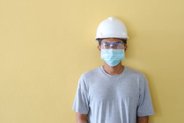 KOVID salgını karşısında güvenlik için kask ve maske takan işçiler sarı bir sahnenin önünde duruyorlar..