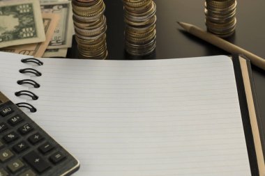 Boş not defteri, kalem, hesap makinesi ve doları banknot, arka planda sikke yığını.